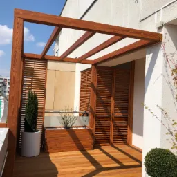 Zadaszony drewnianą konstrukcją taras z deskami, osłonięty drewnianymi panelami żaluzjowymi, z roślinami w donicach, w tle budynki mieszkalne.