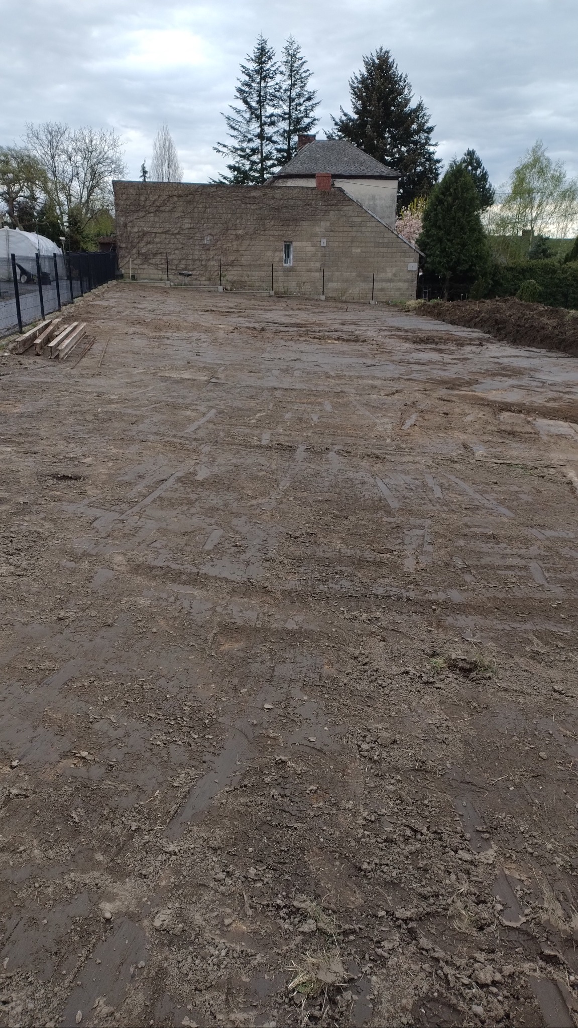 Przygotowany teren pod budowę z widocznymi śladami opon, ogrodzeniem i budynkiem w tle. W rogu leżą drewniane belki.