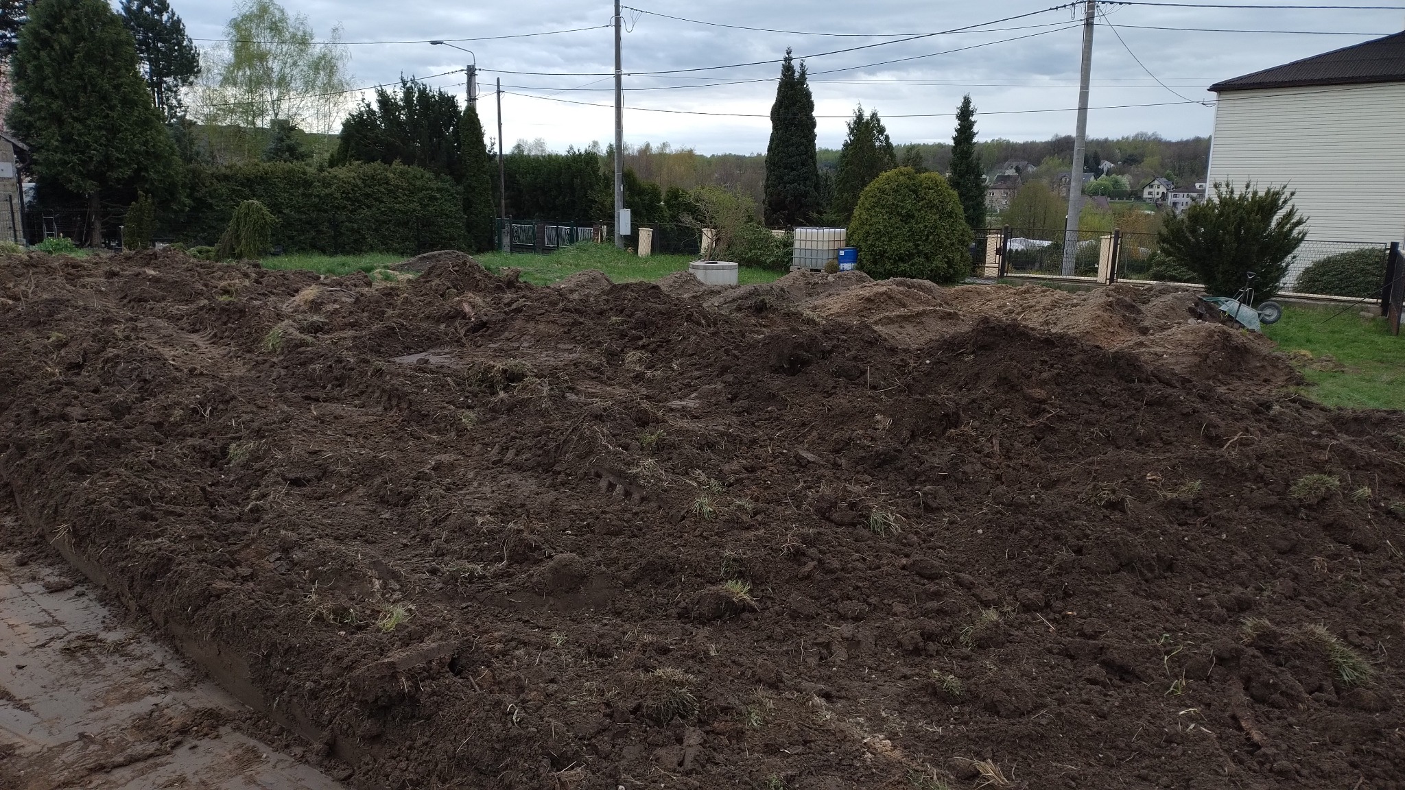 Ukształtowany teren z pryzmami ziemi, widoczne ślady opon. W tle zieleń drzew i zabudowa jednorodzinna, pochmurne niebo. Krajobraz po niwelacji terenu.