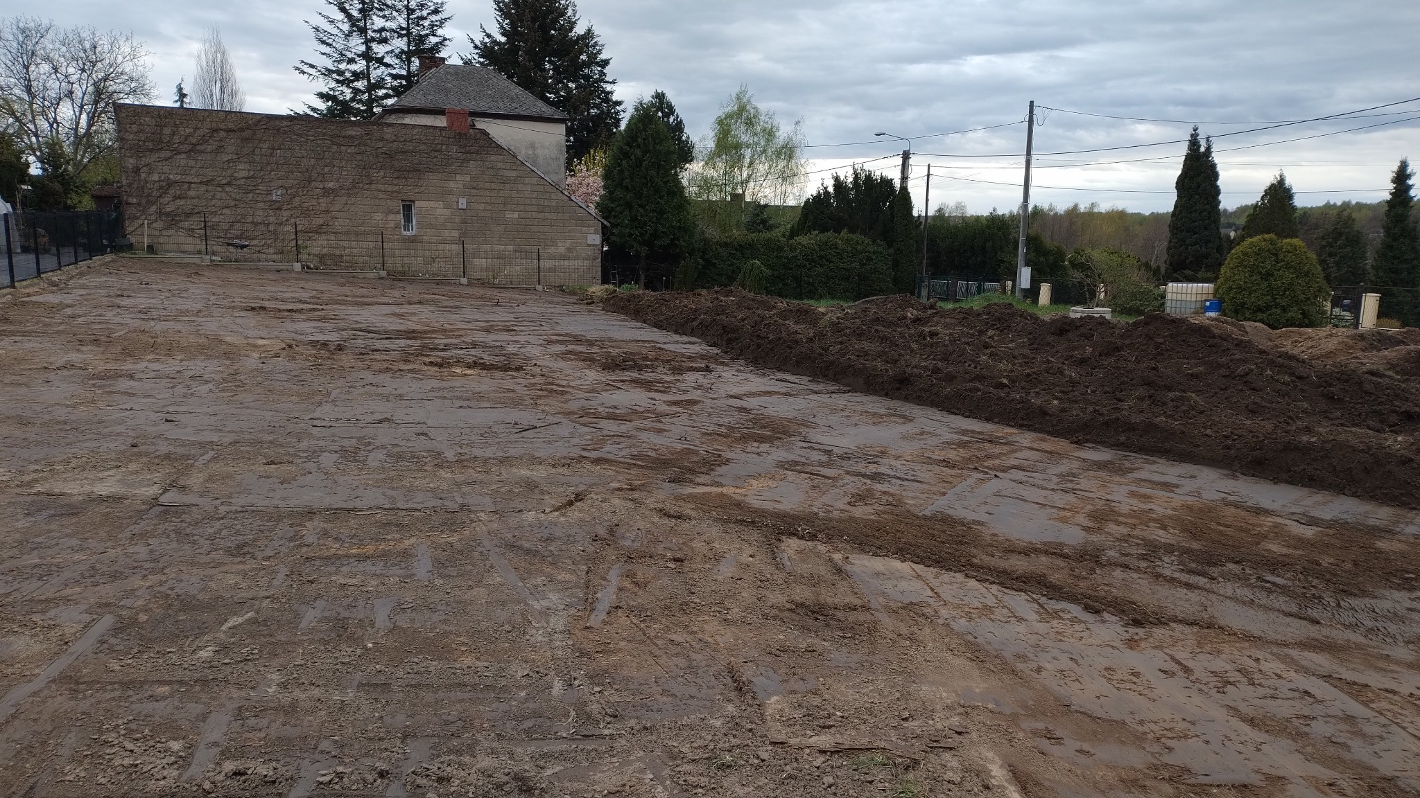 Przygotowany teren pod budowę z widocznymi śladami maszyn, zwały ziemi wzdłuż granicy działki, budynek w tle i pochmurne niebo.