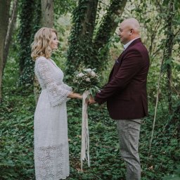 Kociellove - Para trzymająca się za ręce w lesie, kobieta w koronkowej sukience i mężczyzna w bordowej marynarce, bukiet z wstążkami, leśne tło z bluszczem.