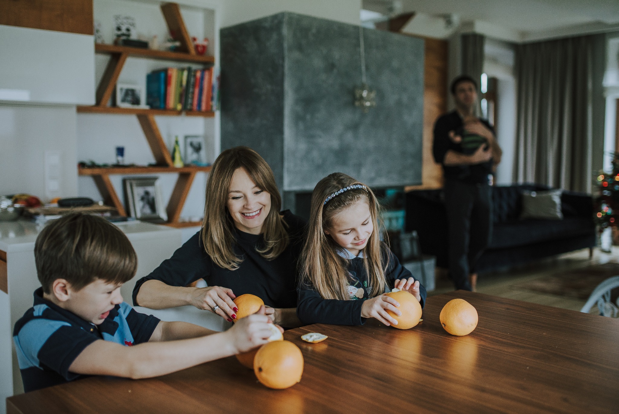 Uśmiechnięta matka z dwójką dzieci obiera pomarańcze przy drewnianym stole w nowoczesnym wnętrzu, widoczny kominek i choinka w tle.