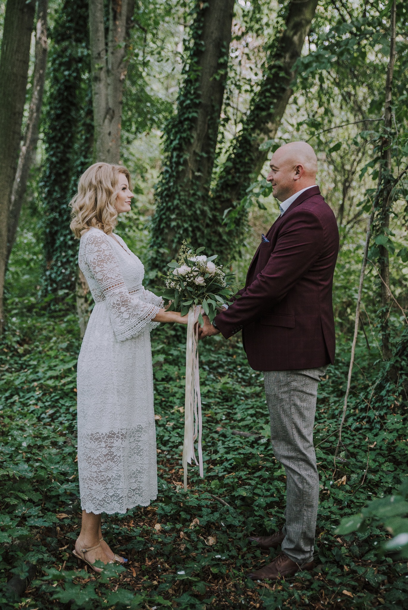 Para trzymająca się za ręce w lesie, kobieta w koronkowej sukience i mężczyzna w bordowej marynarce, bukiet z wstążkami, leśne tło z bluszczem.
