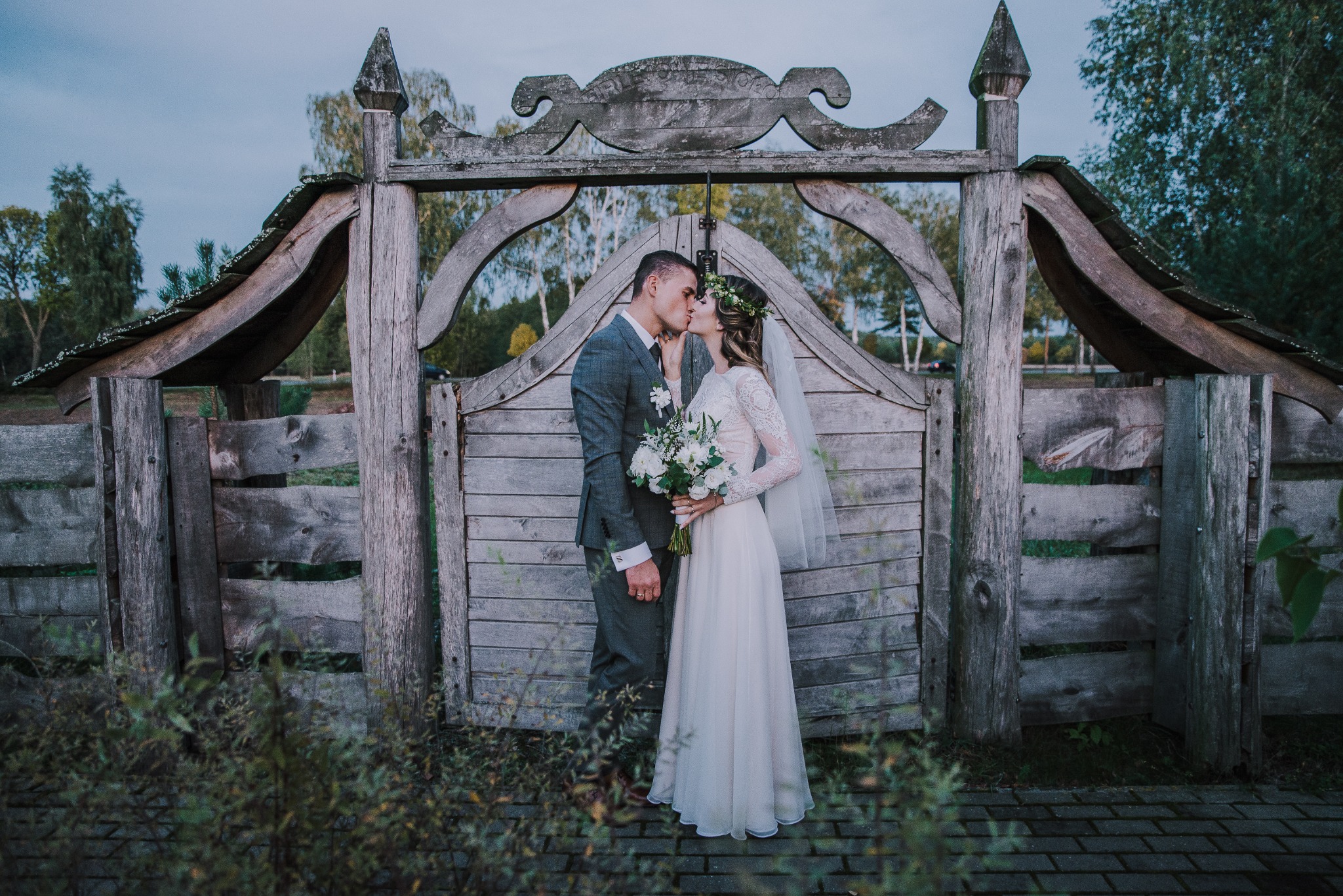 Pocałunek pary młodej na tle rustykalnej drewnianej bramy z dekoracyjnym zwieńczeniem, panna młoda w koronkowej sukni i wianku, pan młody w szarym garniturze.