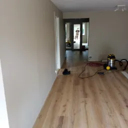 Jasne panele podłogowe w trakcie układania, widoczny odkurzacz przemysłowy, świeżo pomalowane ściany w odcieniach beżu, w tle majster w kombinezonie.