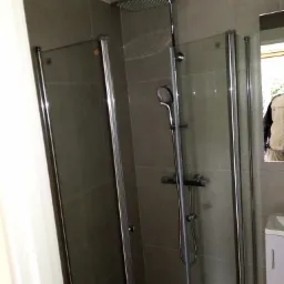 Nowoczesna kabina prysznicowa z deszczownicą i szklanymi drzwiami w łazience wykończonej szarymi płytkami, widoczna umywalka z szafką.