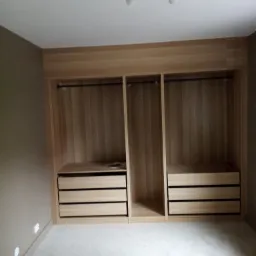 Wnękowa szafa z jasnego drewna z trzema segmentami: dwa z szufladami i drążkami na ubrania, jeden pusty, oświetlenie sufitowe z trzema reflektorami, ściana w kolorze taupe, jasna podłoga.