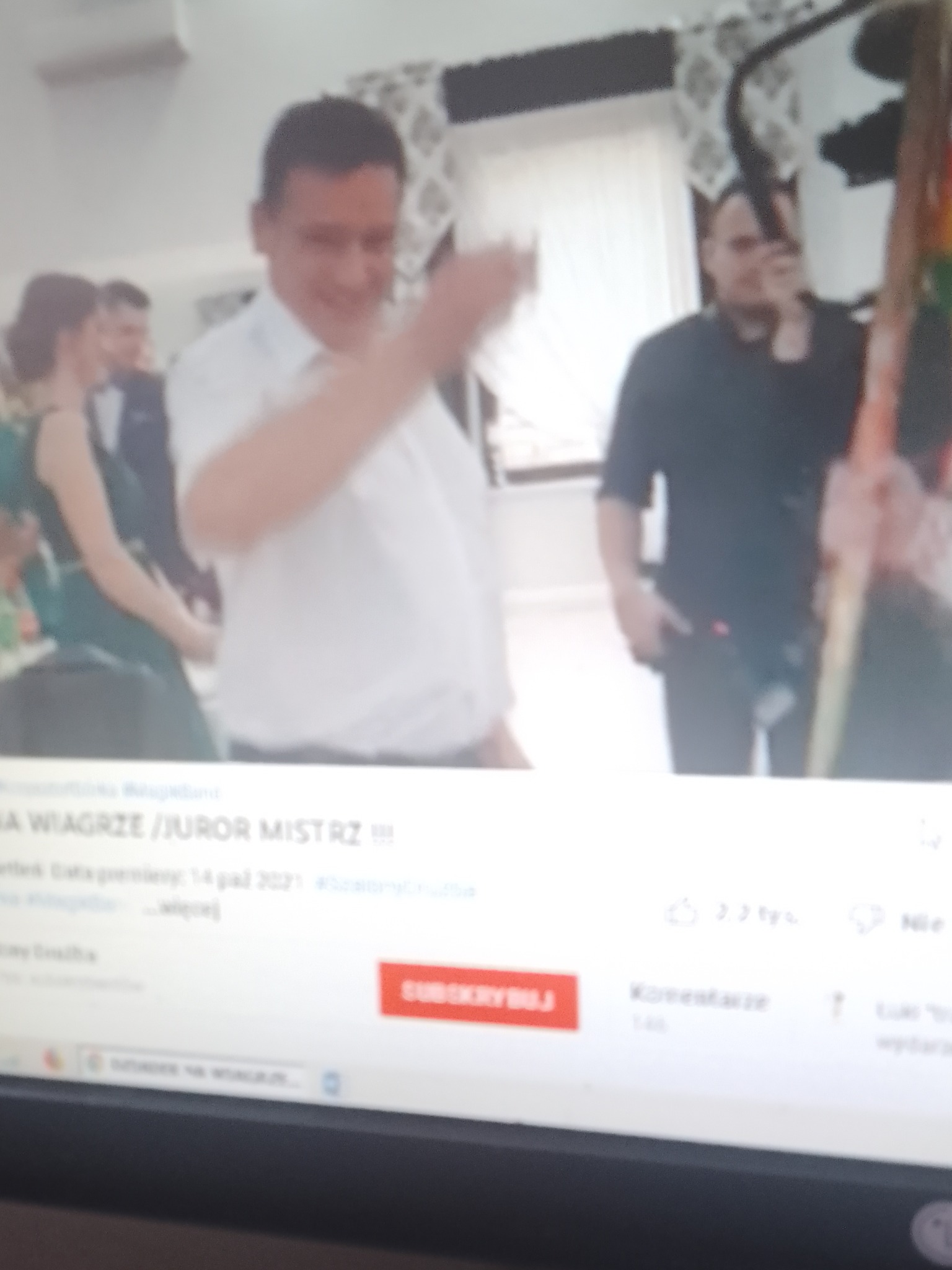 Kadr z nagrania wideo przedstawiający mężczyznę w białej koszuli gestykulującego, prawdopodobnie podczas wydarzenia lub konkursu. W tle widoczni inni ludzie i elementy wystroju wnętrza.