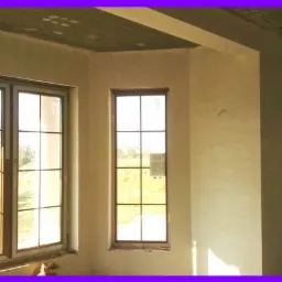 Wnętrze w stanie surowym z nałożonymi tynkami gipsowymi, widoczne trzy okna z ciemnymi ramami i podziałami, sufit z widocznym betonem i śladami po szalunkach.