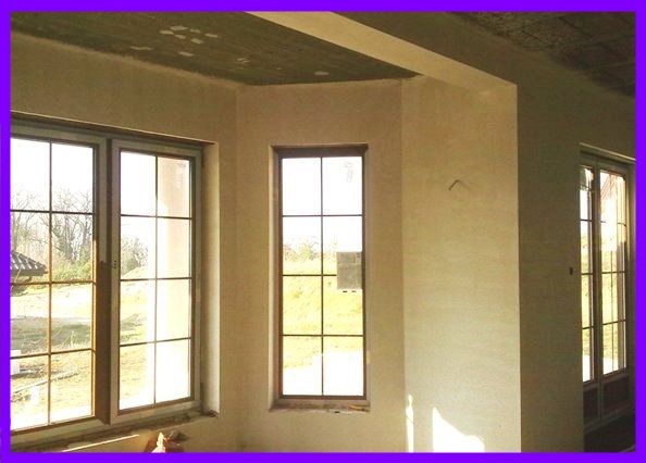 Wnętrze w stanie surowym z nałożonymi tynkami gipsowymi, widoczne trzy okna z ciemnymi ramami i podziałami, sufit z widocznym betonem i śladami po szalunkach.
