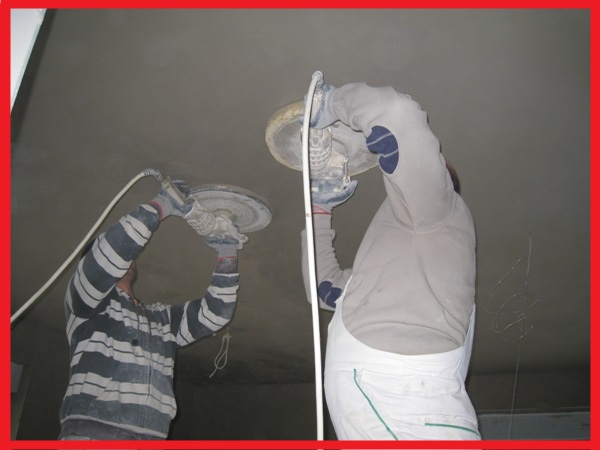 Dwaj pracownicy w trakcie szlifowania sufitu po nałożeniu tynku cementowo-wapiennego maszynowego, używają szlifierek z okrągłymi tarczami, widoczne kable zasilające i ślady tynku na suficie.