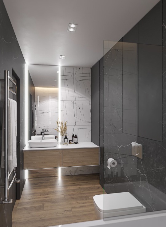 Nowoczesna łazienka z marmurowymi ścianami, drewnianą szafką pod umywalką, podświetlanym lustrem i widoczną częścią toalety oddzielonej szklaną ścianką.