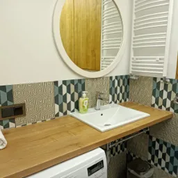 Łazienka z drewnianym blatem, białą umywalką, okrągłym lustrem i grzejnikiem drabinkowym, dekoracyjne płytki na ścianie, pralka pod blatem.