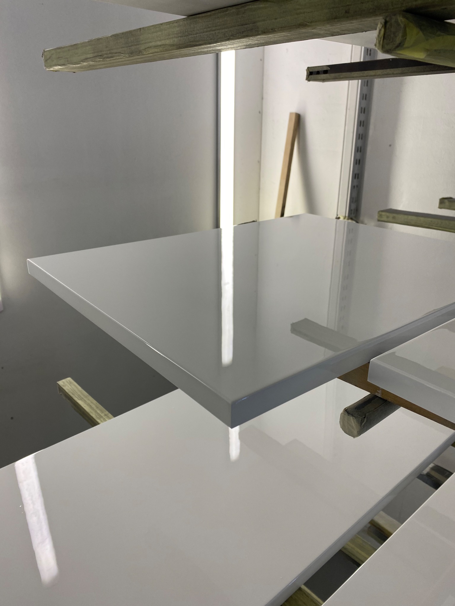 Białe, lakierowane elementy meblowe na drewnianych stojakach w lakierni, widoczne odbicia światła na powierzchni.