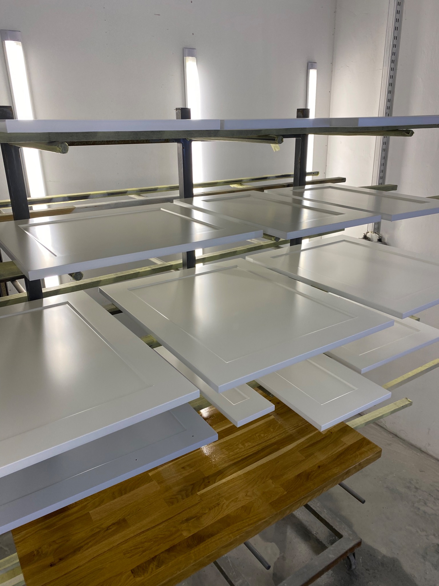 Białe, świeżo polakierowane fronty meblowe schnące na metalowym regale w warsztacie, z widocznym drewnianym blatem roboczym poniżej.