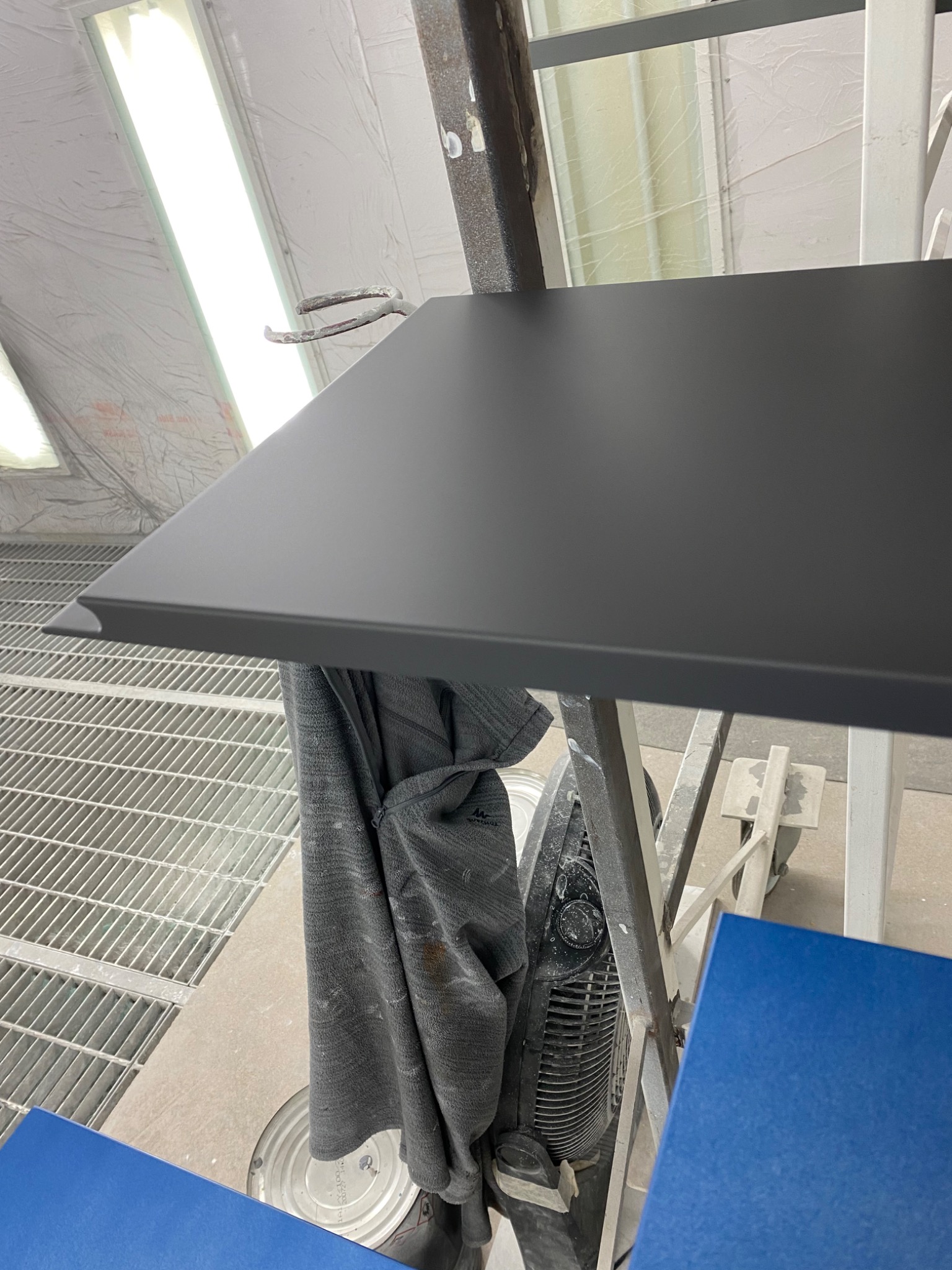 Czarny blat z zaokrąglonym brzegiem, prawdopodobnie świeżo lakierowany, w pomieszczeniu lakierniczym z kratą podłogową i widocznym wentylatorem oraz fragmentem niebieskiej płyty.
