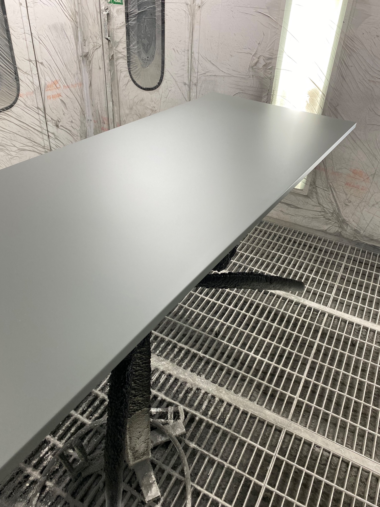 Pomalowany na szaro blat, prawdopodobnie drewniany, suszony w kabinie lakierniczej, widoczna kratka pod blatem i zacieki farby na stojaku.