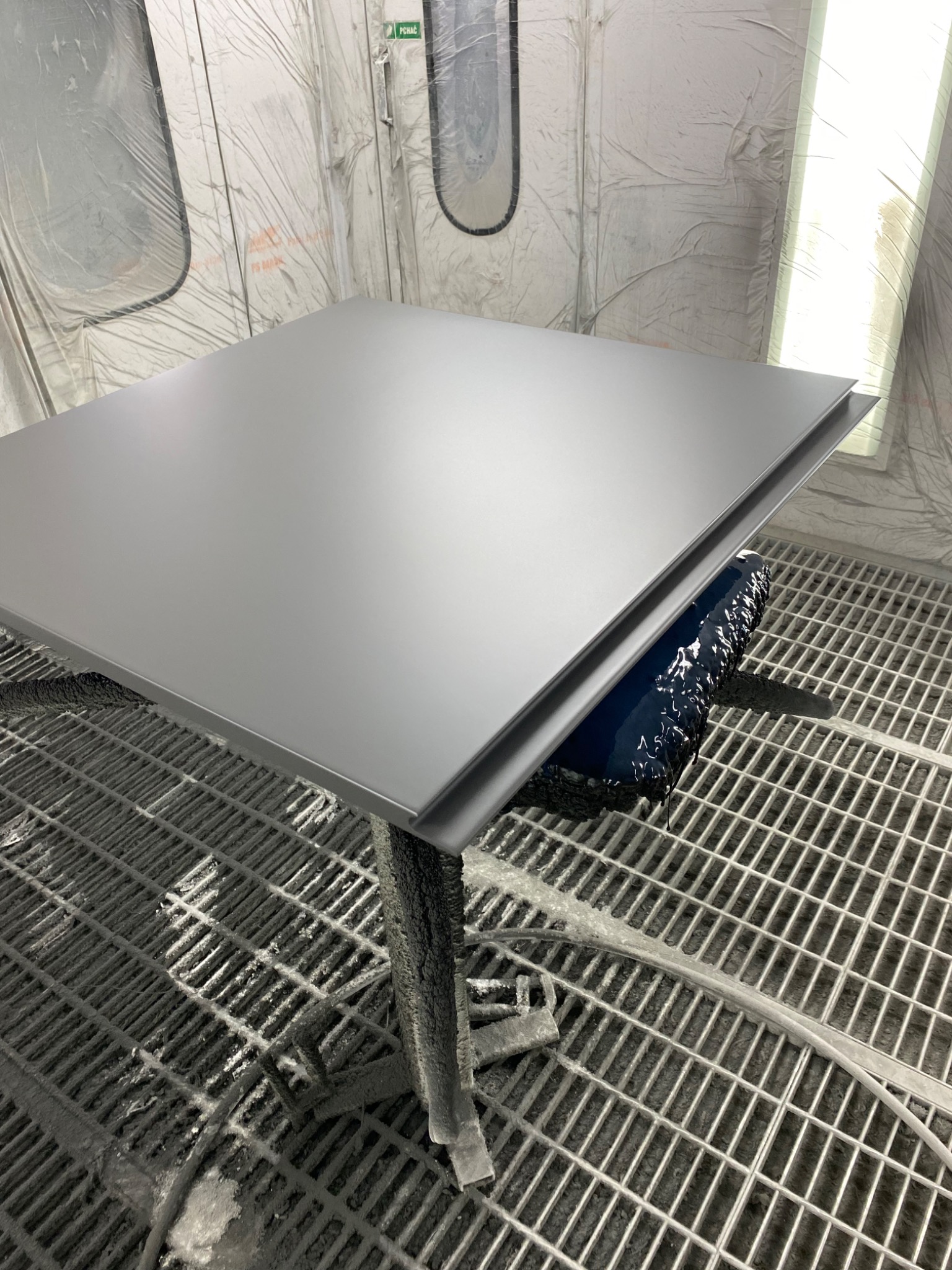 Szary blat stołu z metalowym obramowaniem, świeżo po lakierowaniu, ustawiony na czarnym podkładzie w komorze lakierniczej.