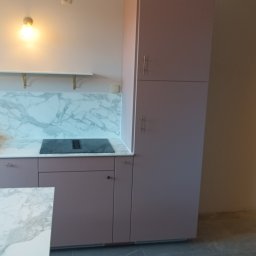 Dan-fliz firma ogólnobudowlana - Nowoczesna kuchnia z szafkami w kolorze pudrowego różu, marmurowym blatem i panelem ściennym, czarną płytą indukcyjną z okapem oraz ciemnoszarą podłogą. Nad blatem biała półka ze złotymi wspornikami.