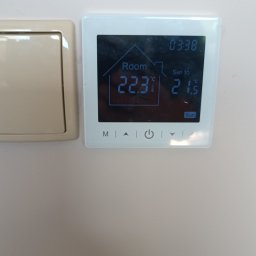 Dan-fliz firma ogólnobudowlana - Biały cyfrowy termostat ścienny z wyświetlaczem pokazującym temperaturę pokojową 22.3°C i ustawioną 21.5°C, obok beżowy włącznik światła na jasnej ścianie.