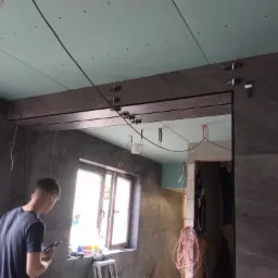 Montaż płyty karton gips na suficie w trakcie remontu łazienki z ciemnymi płytkami, widoczny pracownik z telefonem i narzędzia na parapecie.