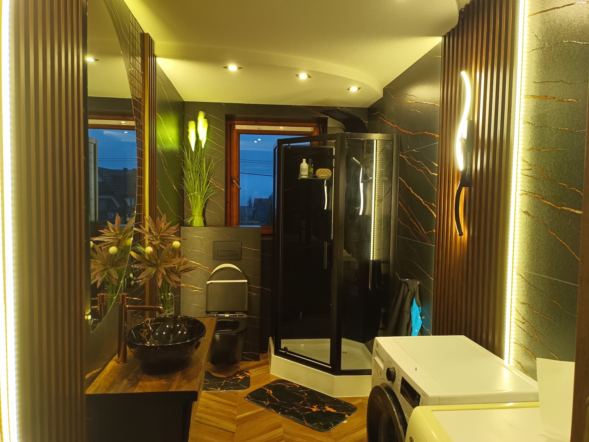 Nowoczesna łazienka z czarną armaturą i marmurowymi płytkami, oświetlona listwami LED i punktowymi lampami. Uwagę przykuwa deska z drewna egzotycznego pod umywalką.