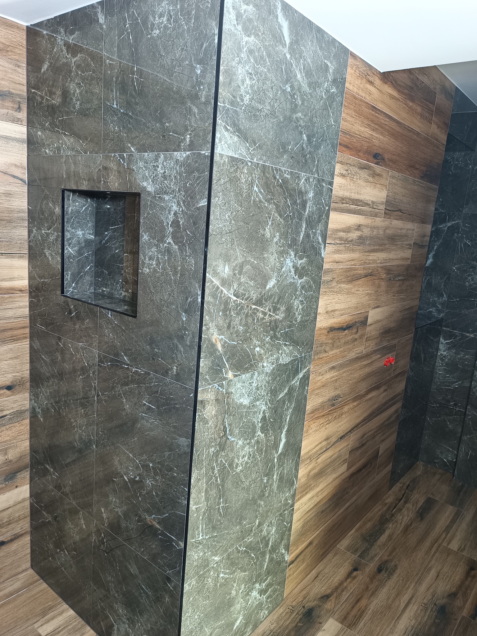 Nowoczesna łazienka z ciemnymi, marmurkowymi płytkami i wnęką w ścianie. Ułożenie płytek imituje drewno, tworząc ciepły kontrast.