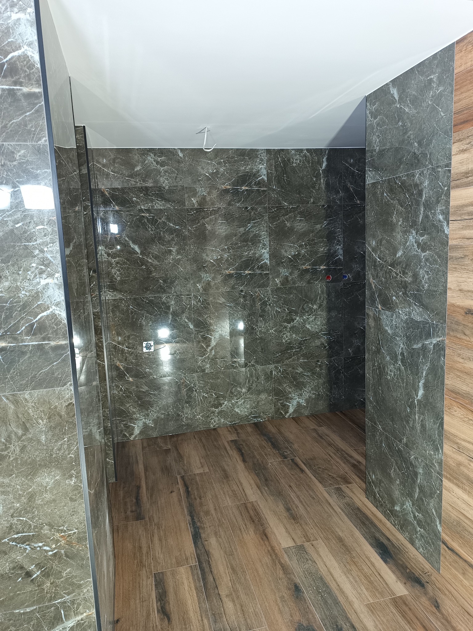 Wykończona łazienka z płytkami imitującymi marmur na ścianach i drewnopodobną podłogą. Widoczne przyłącza wodne i sufit z wystającym kablem.