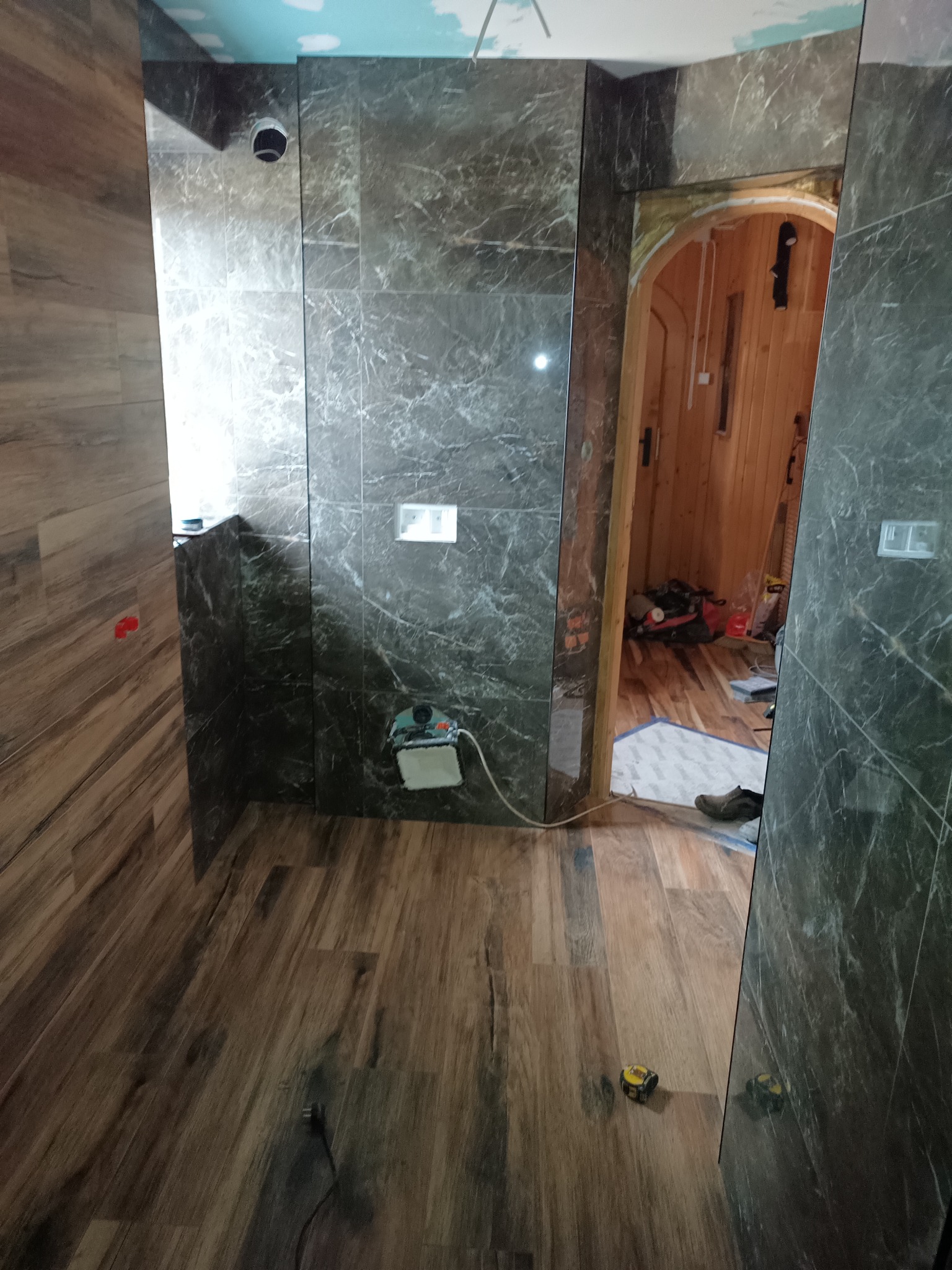 Łazienka w trakcie wykańczania: marmurowe płytki na ścianach, drewnopodobna podłoga. Widoczne narzędzia i instalacja. Sufit z motywem nieba.