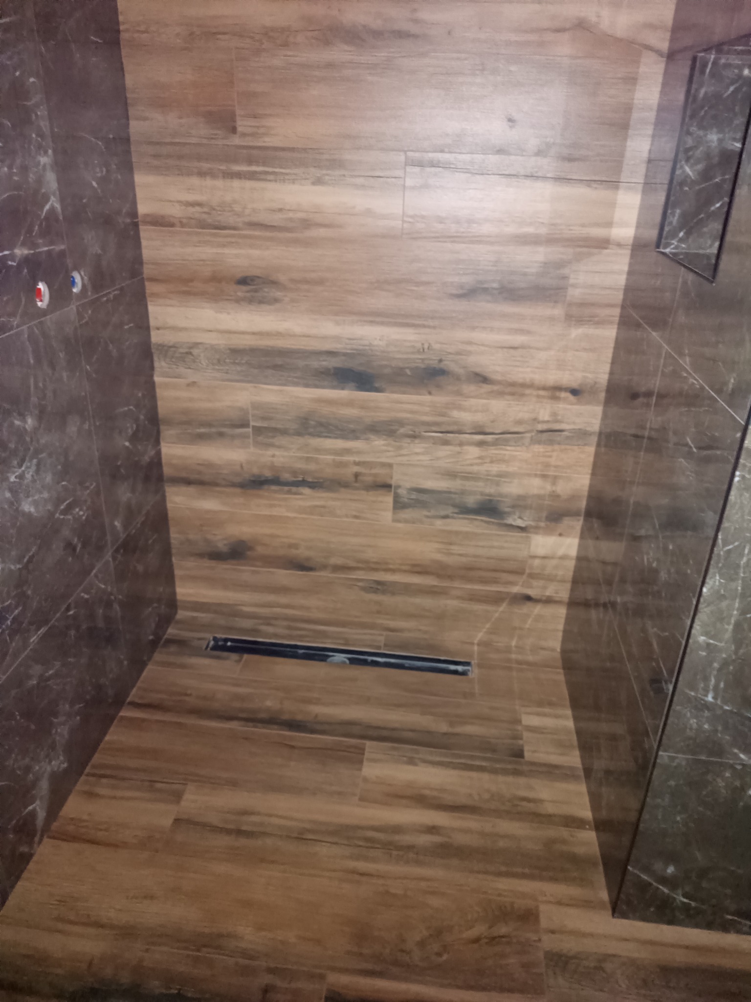 Ukończona kabina prysznicowa z odpływem liniowym, wyłożona płytkami imitującymi drewno na podłodze i marmur na ścianach, z widocznymi zaworami.