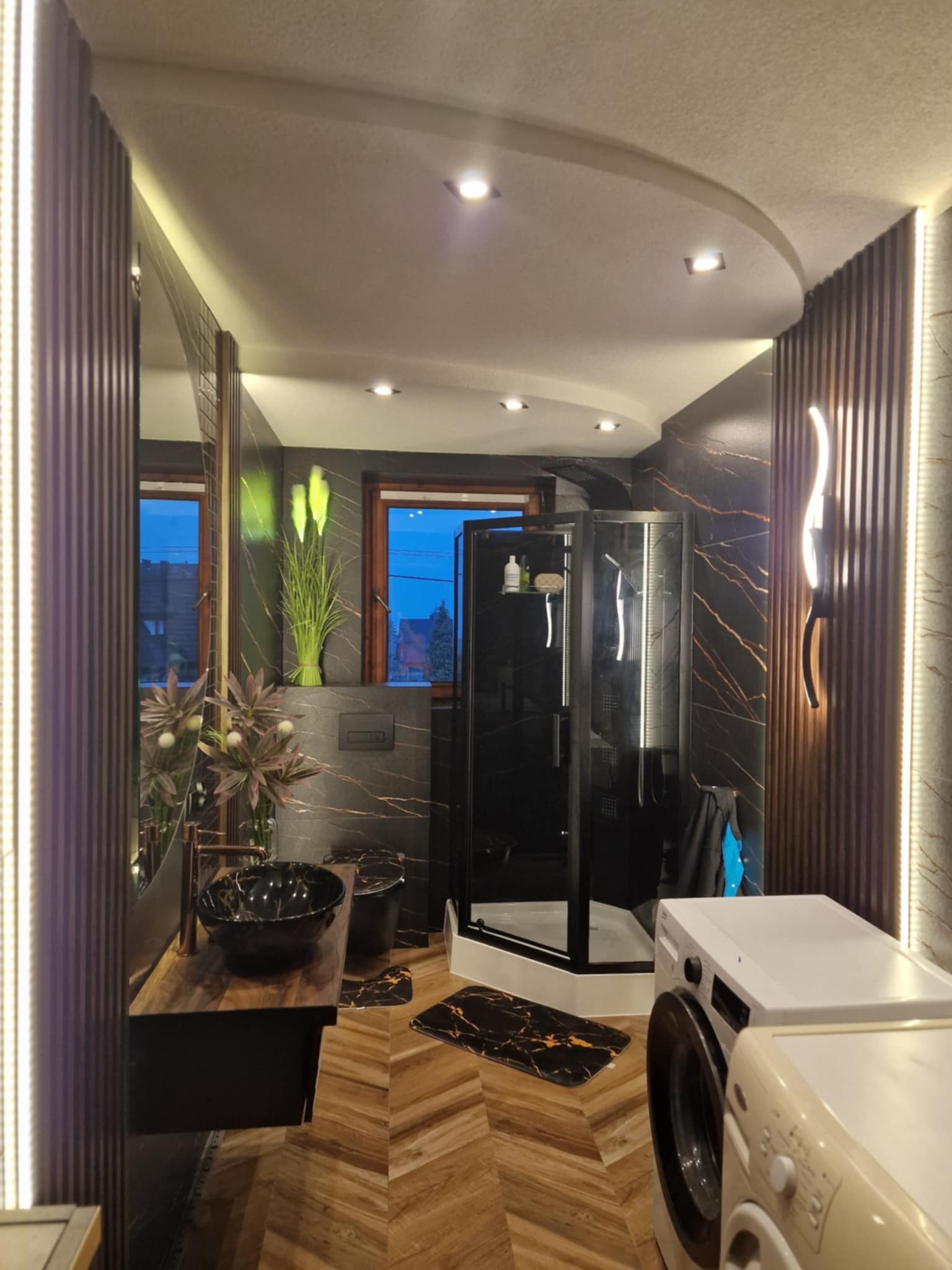 Nowoczesna łazienka z czarną kabiną prysznicową, umywalką w marmurkowe wzory i drewnianą podłogą. Oświetlenie LED podkreśla detale. Widoczne dwie pralki.