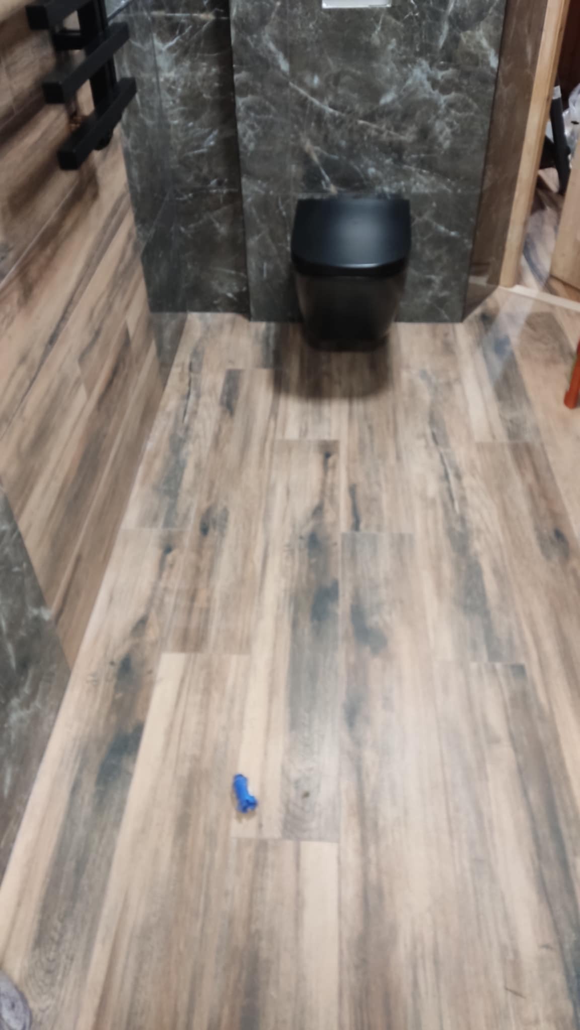 Wykończona łazienka z podwieszaną miską WC, płytkami imitującymi drewno na podłodze i ścianie. Na podłodze leży niebieski element montażowy.