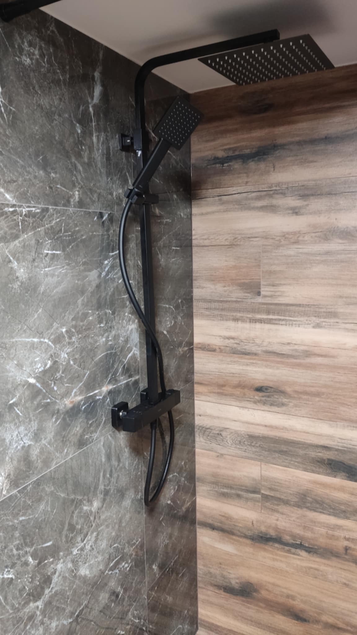 Czarny zestaw prysznicowy z deszczownicą, zamontowany na ścianie wyłożonej szarymi płytkami imitującymi marmur i drewnopodobnymi, brązowymi płytkami.
