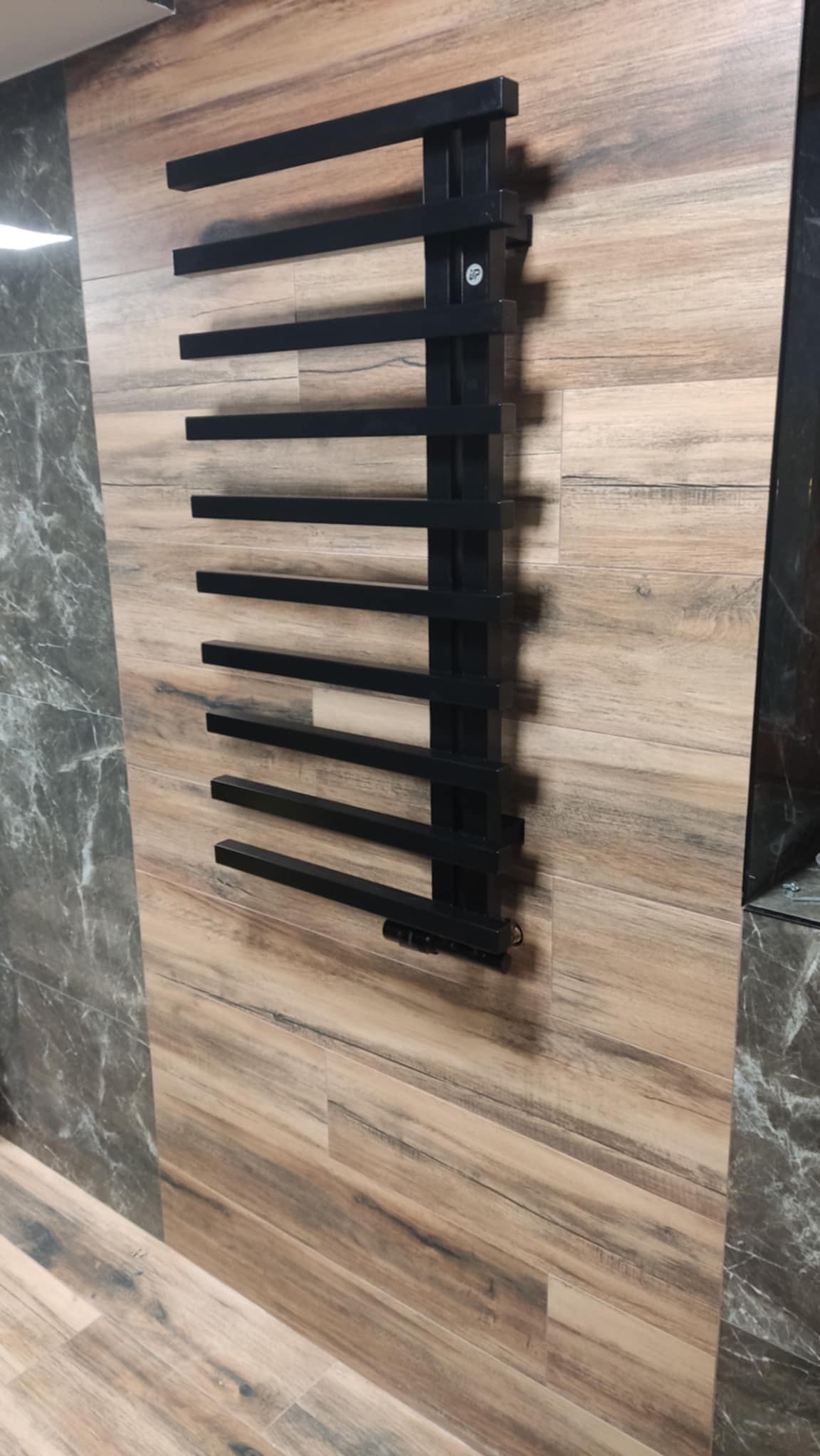 Czarny, nowoczesny grzejnik łazienkowy zamontowany na ścianie wyłożonej płytkami imitującymi drewno. Widoczny fragment marmurowej ściany.
