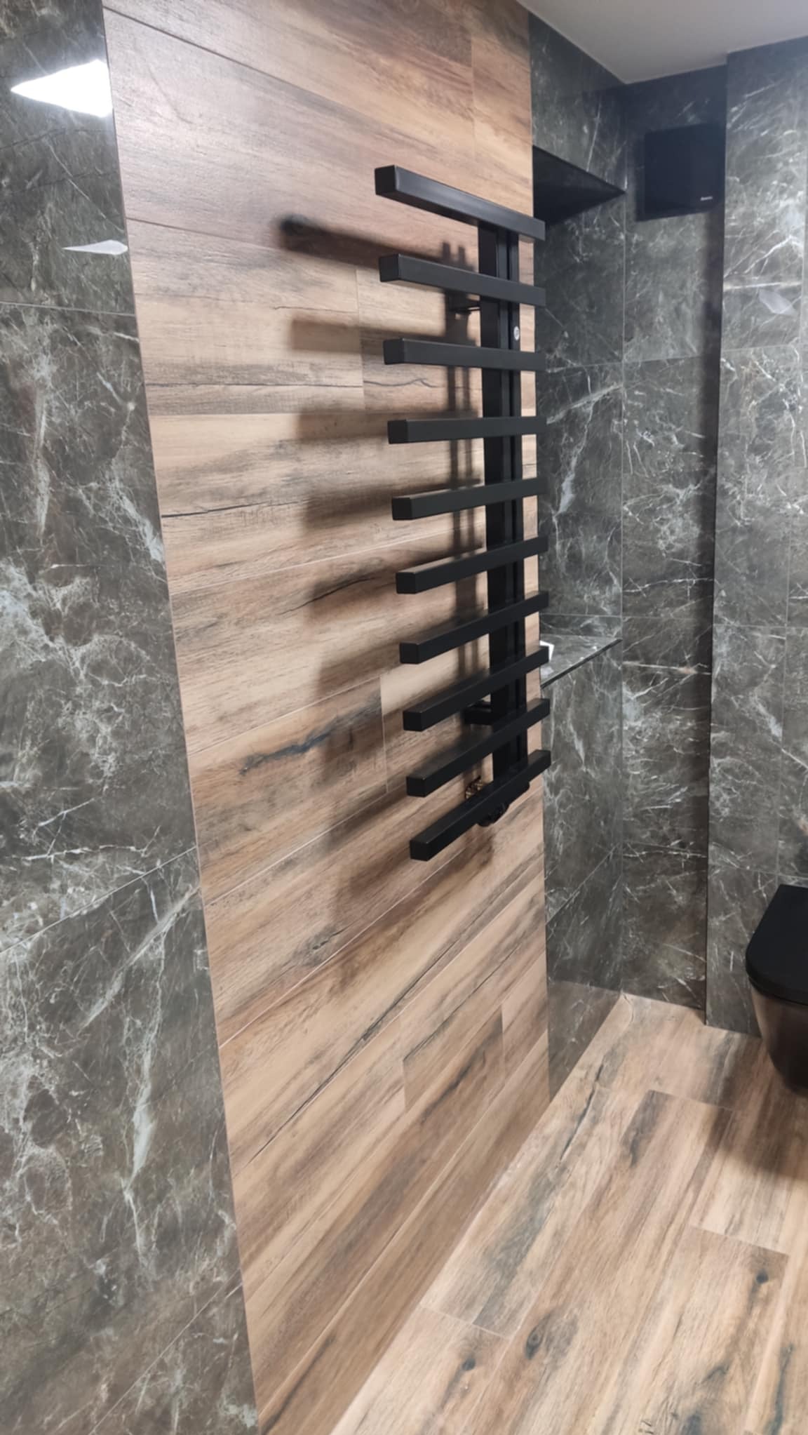 Wykończona łazienka z płytkami imitującymi drewno i marmur, z czarnym grzejnikiem drabinkowym. Kontrast materiałów i kolorów w nowoczesnym wnętrzu.