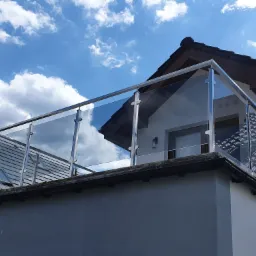 Szklana balustrada balkonowa z elementami ze stali nierdzewnej, widok z dołu na tle błękitnego nieba z chmurami, mocowana do ściany budynku.