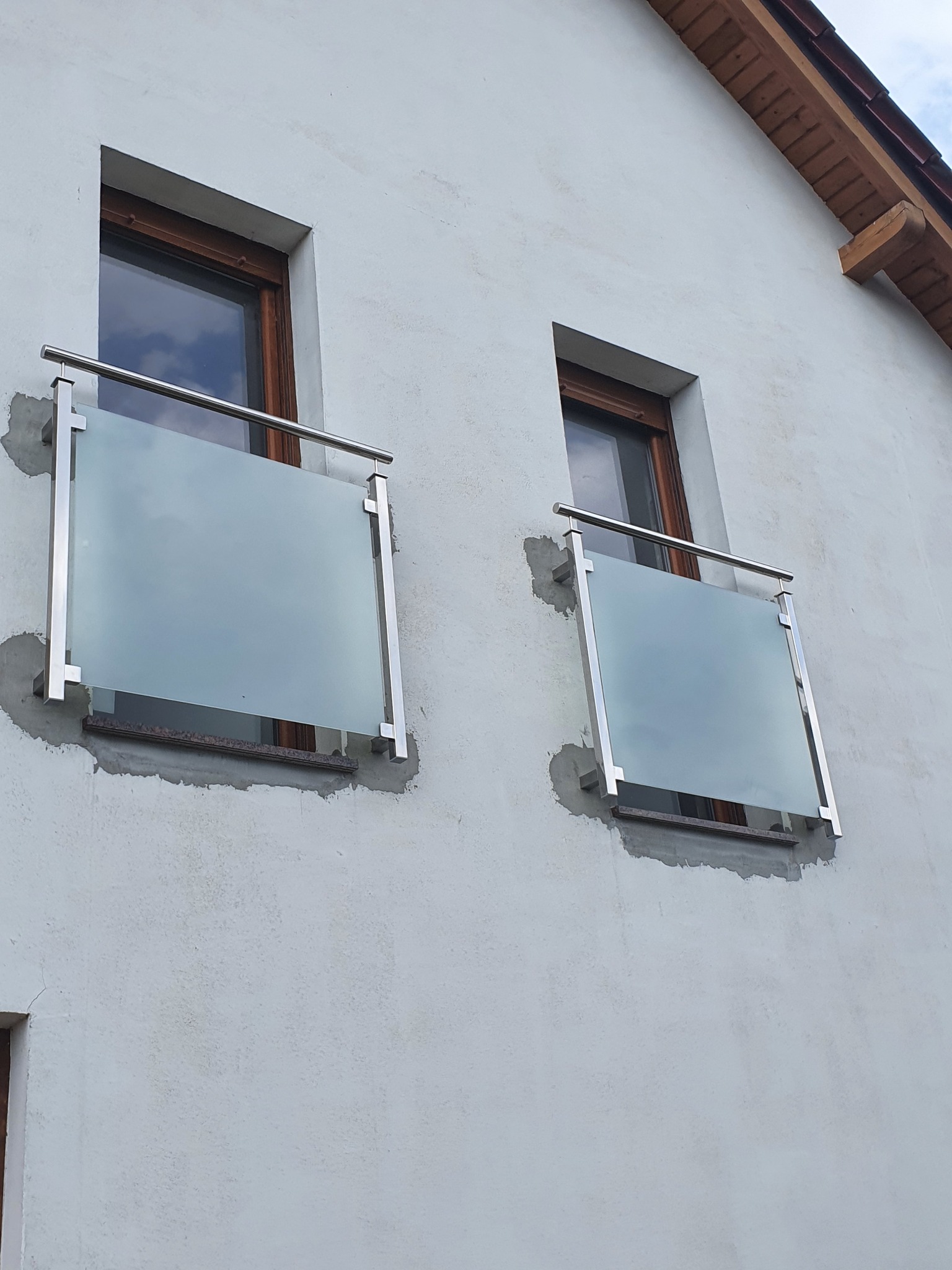 Dwa okna z zamontowanymi nowoczesnymi, szklanymi balustradami z elementami ze stali nierdzewnej na elewacji domu, widoczne ślady po montażu.