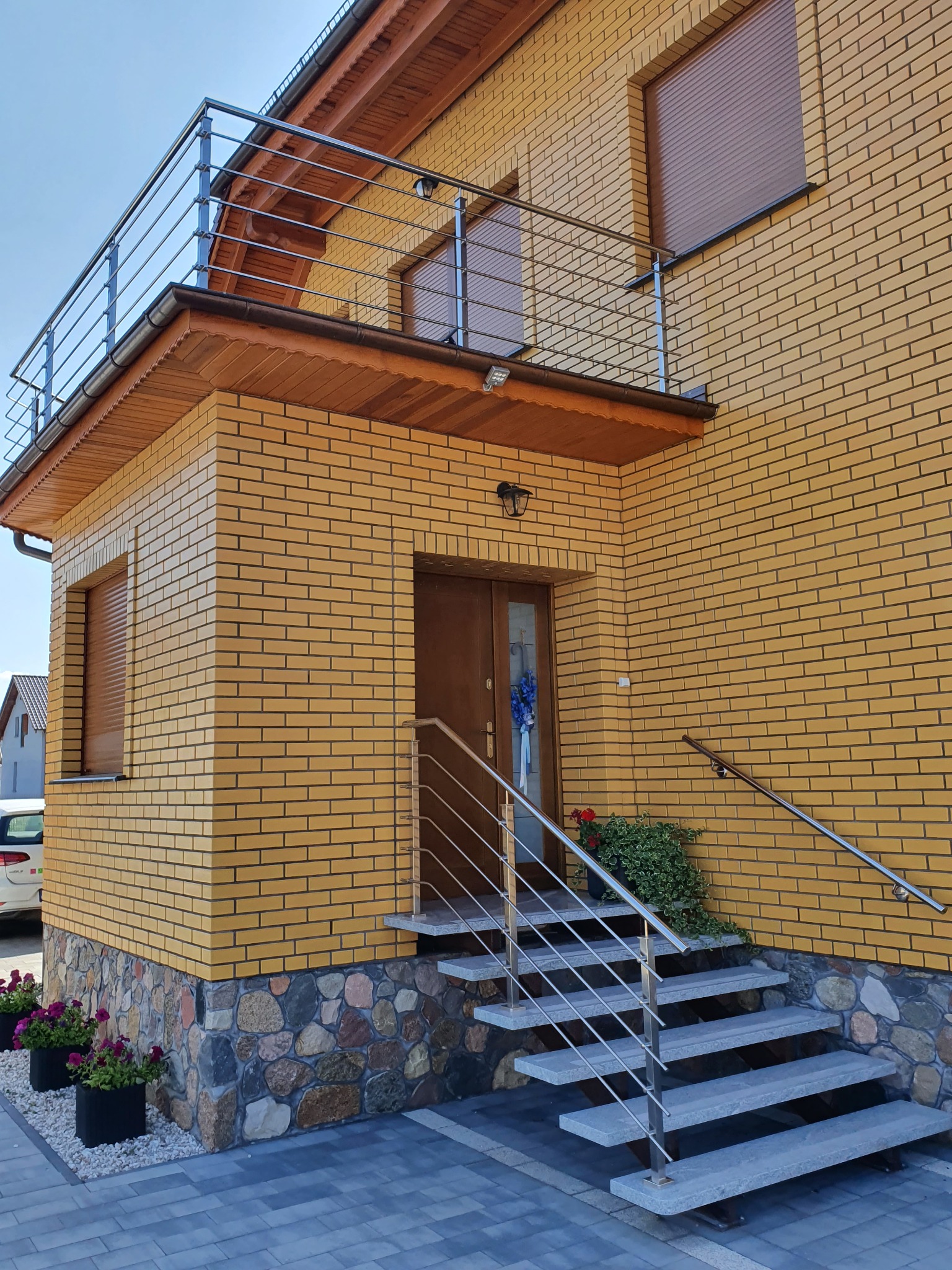 Dom z żółtej cegły z balkonem i schodami z balustradami ze stali nierdzewnej oraz kamiennym podłożem, widoczne kwiaty w donicach.