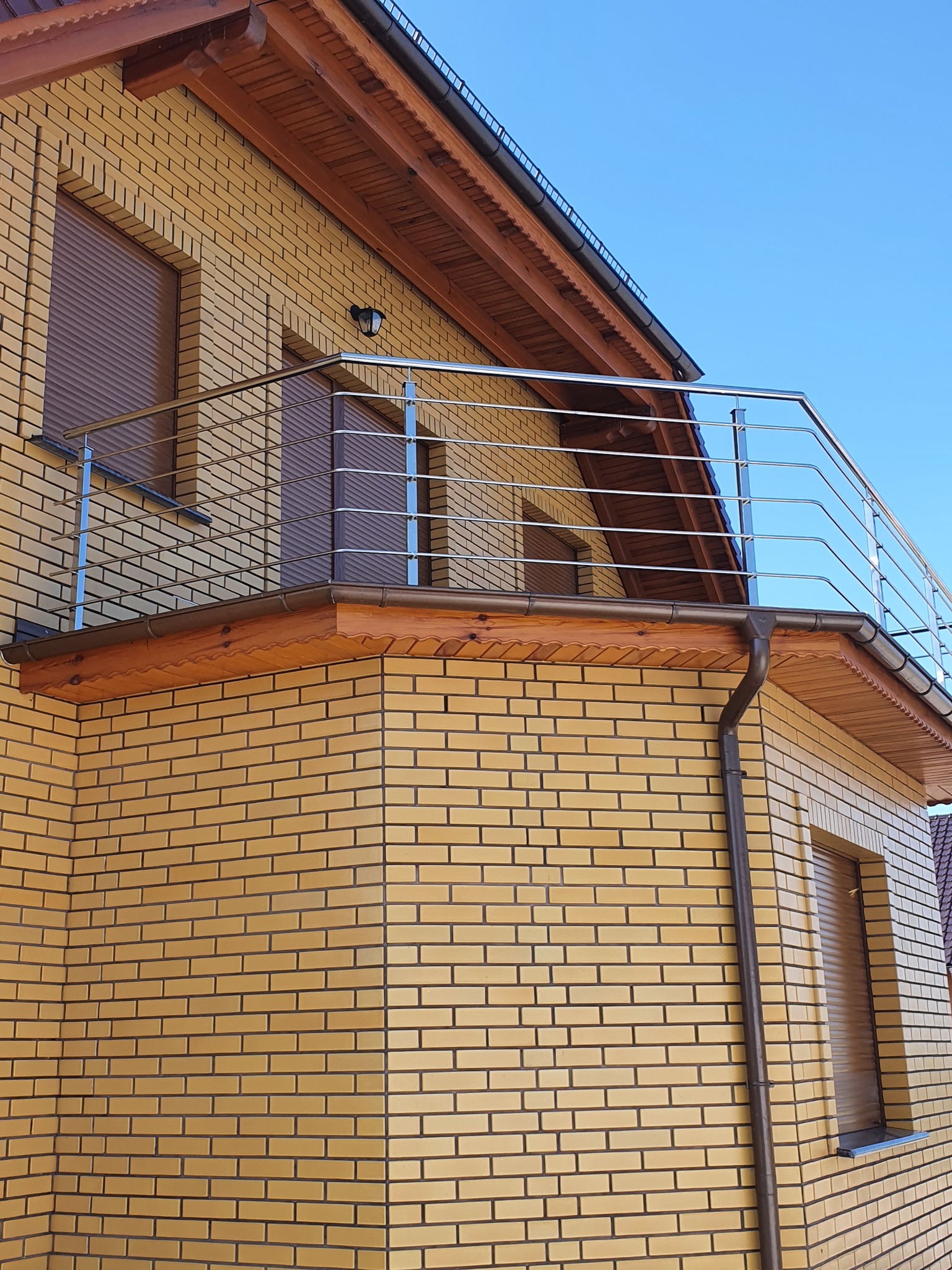 Balkon z nowoczesną, stalową balustradą na tle żółtej elewacji budynku z cegły klinkierowej i brązowymi okiennicami.