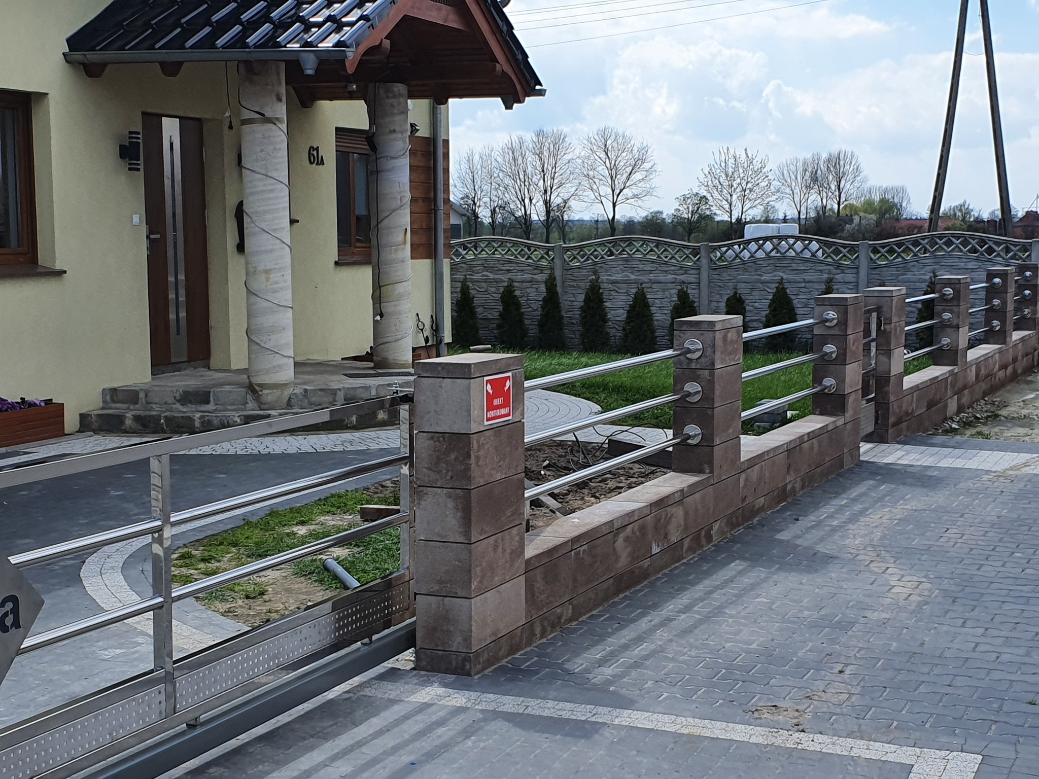 Nowoczesna posesja z przesuwną bramą i ogrodzeniem z elementami murowanymi oraz poziomymi rurami ze stali nierdzewnej, widok na dom z beżową elewacją i kolumnami.