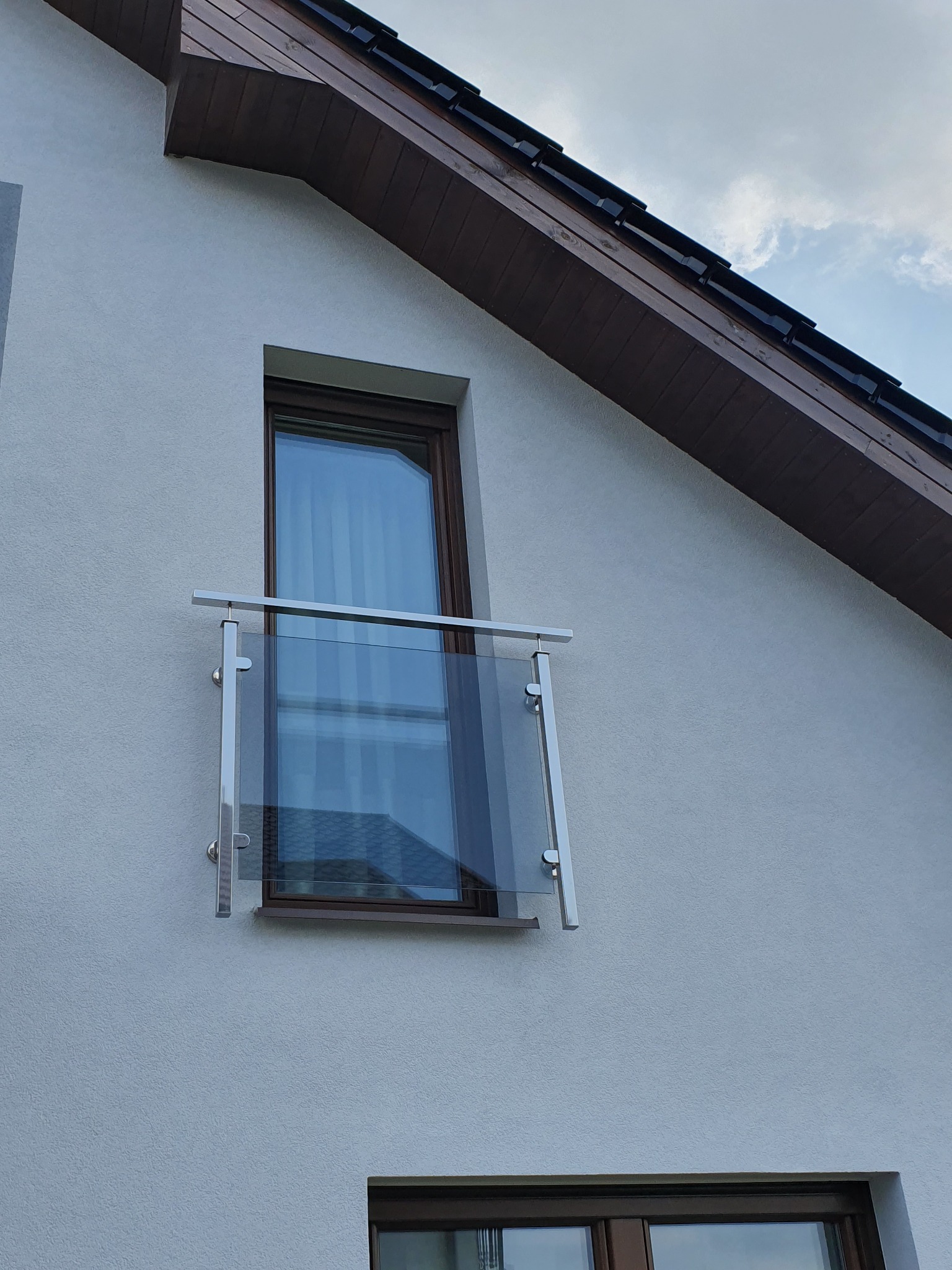 Szklana balustrada balkonowa z elementami ze stali nierdzewnej zamontowana przy oknie na elewacji budynku z brązowym dachem.