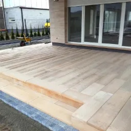 Jasna, drewniana podłoga tarasu, wykonana z desek imitujących kostkę, z niskimi schodami i obrzeżem z szarej kostki, widok z poziomu gruntu, w tle budynek mieszkalny i żółty samochód dostawczy.