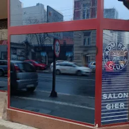 Okno wystawowe z czerwoną ramą, odbijające ruch uliczny i budynki, z widocznym napisem 'ADMIRAŁ CLUB SALON GIER' na szybie.