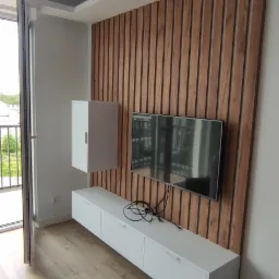 Nowoczesny salon z drewnianą ścianą z lameli, wiszącą białą szafką RTV i telewizorem, widok na balkon.