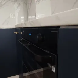 Fragment nowoczesnej kuchni z ciemnoniebieskimi szafkami z poziomymi, złotymi uchwytami i wbudowanym piekarnikiem z wyświetlaczem, na blacie z białego marmuru.