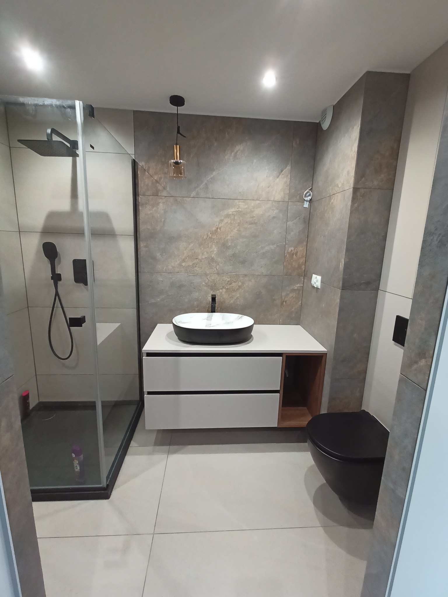 Nowoczesna łazienka z kabiną prysznicową typu walk-in, umywalką nablatową na szafce z szufladami, podwieszaną miską WC w kolorze czarnym i płytkami imitującymi kamień na ścianach.