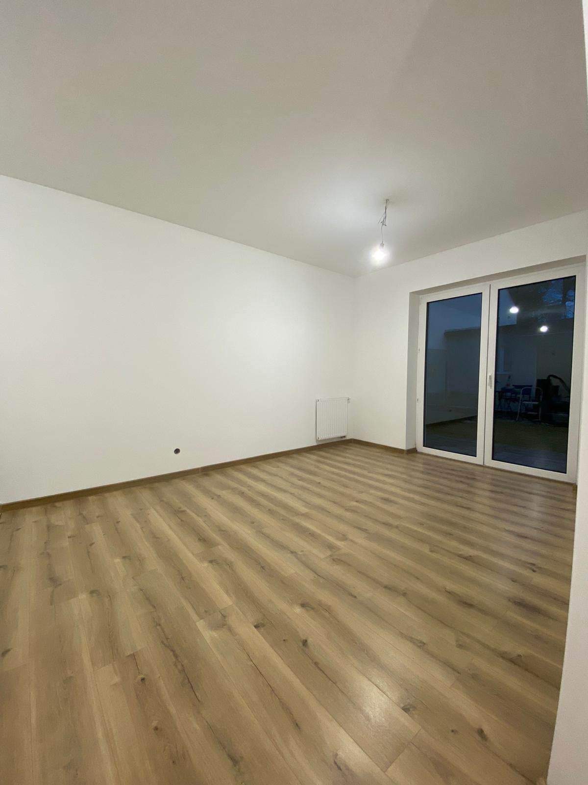 Pusty pokój po remoncie z panelami podłogowymi w odcieniach drewna, świeżo pomalowanymi białymi ścianami i dużymi przesuwnymi drzwiami balkonowymi.