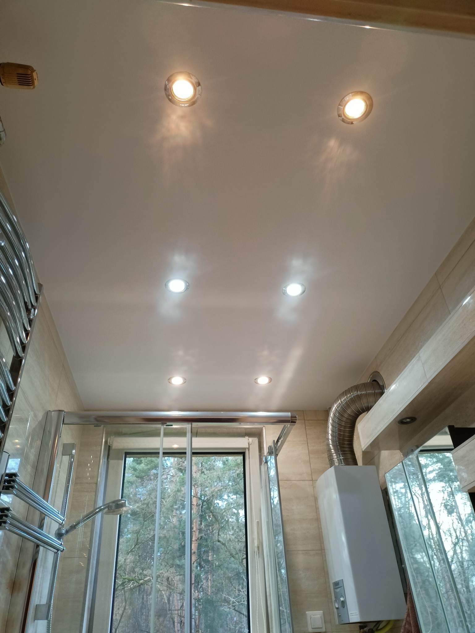 Biały sufit podwieszany z wbudowanymi reflektorami w łazience, widoczny bojler z rurą odprowadzającą spaliny oraz fragment kabiny prysznicowej z widokiem na las za oknem.