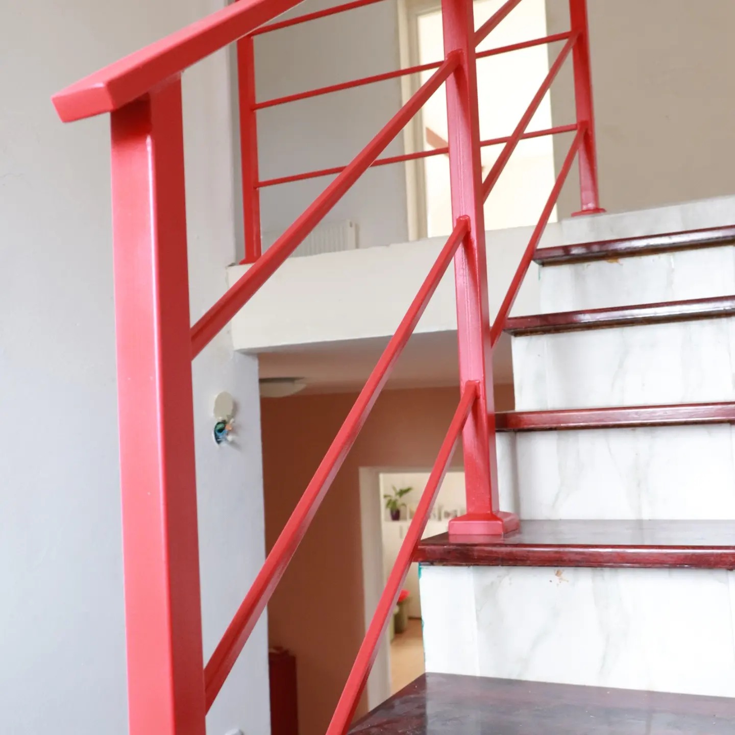 Czerwona, metalowa balustrada schodowa z poziomymi prętami, marmurowe stopnie z ciemnobrązowymi krawędziami, widok na wnętrze domu w tle.
