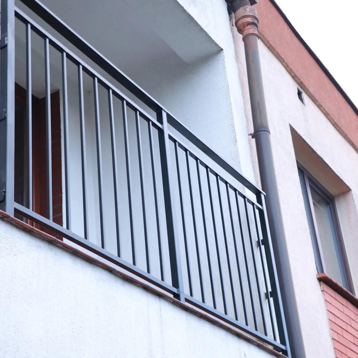 Czarna, metalowa balustrada balkonowa z pionowymi szczebelkami, zamontowana na balkonie budynku z białą elewacją i brązową rynną spustową.